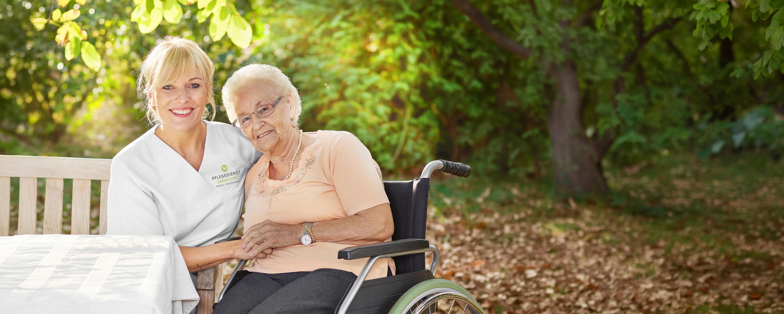 Pflegekraft des Pflegedienst Zwönitztal sitzt neben einer lächelnden Seniorin im Rollstuhl auf einer Gartenbank und hält ihre Hand